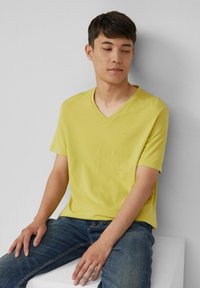 T-shirt jaune en V fait en tissu doux, à manches courtes, avec un logo subtil. Le modèle porte un jean bleu, assis sur une surface blanche.