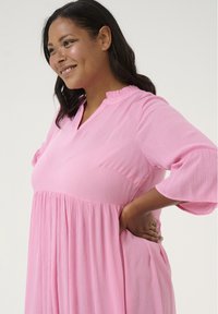 Robe rose ample avec un col volanté et des manches cloche 3/4. Le tissu présente un motif texturé subtil et une silhouette fluide.