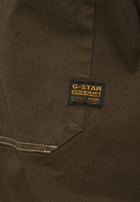 Brunt tyg med en liten svart och orange G-Star Originals Raw Denim märkning sydd nära en beige dubbelsydd söm.