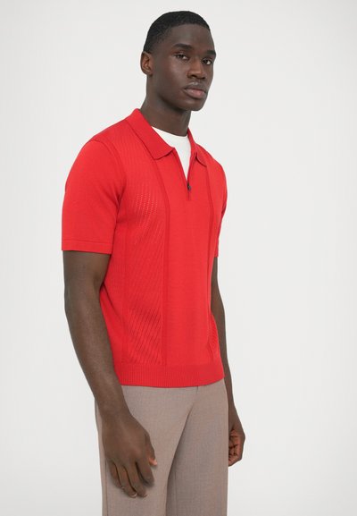 Punainen lyhythihainen poolopaita, jossa on teksturoitu kuvio sivuilla ja vetoketjullinen kaulus. Päällä vaalean beiget housut.