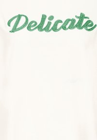 Chemise blanche à manches courtes avec texte brodé vert « Delicate », texture lisse et design décontracté.