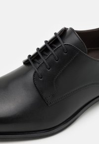 Schwarze Lederdressschuhe mit glatter Oberfläche, sechs Ösen, geschwungener Nahtverarbeitung und einem leicht erhöhten Absatz.