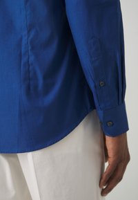 Manica e polsino di una camicia a maniche lunghe blu con bottoni neri, indossata da una persona in pantaloni bianchi, con la mano rilassata al lato.
