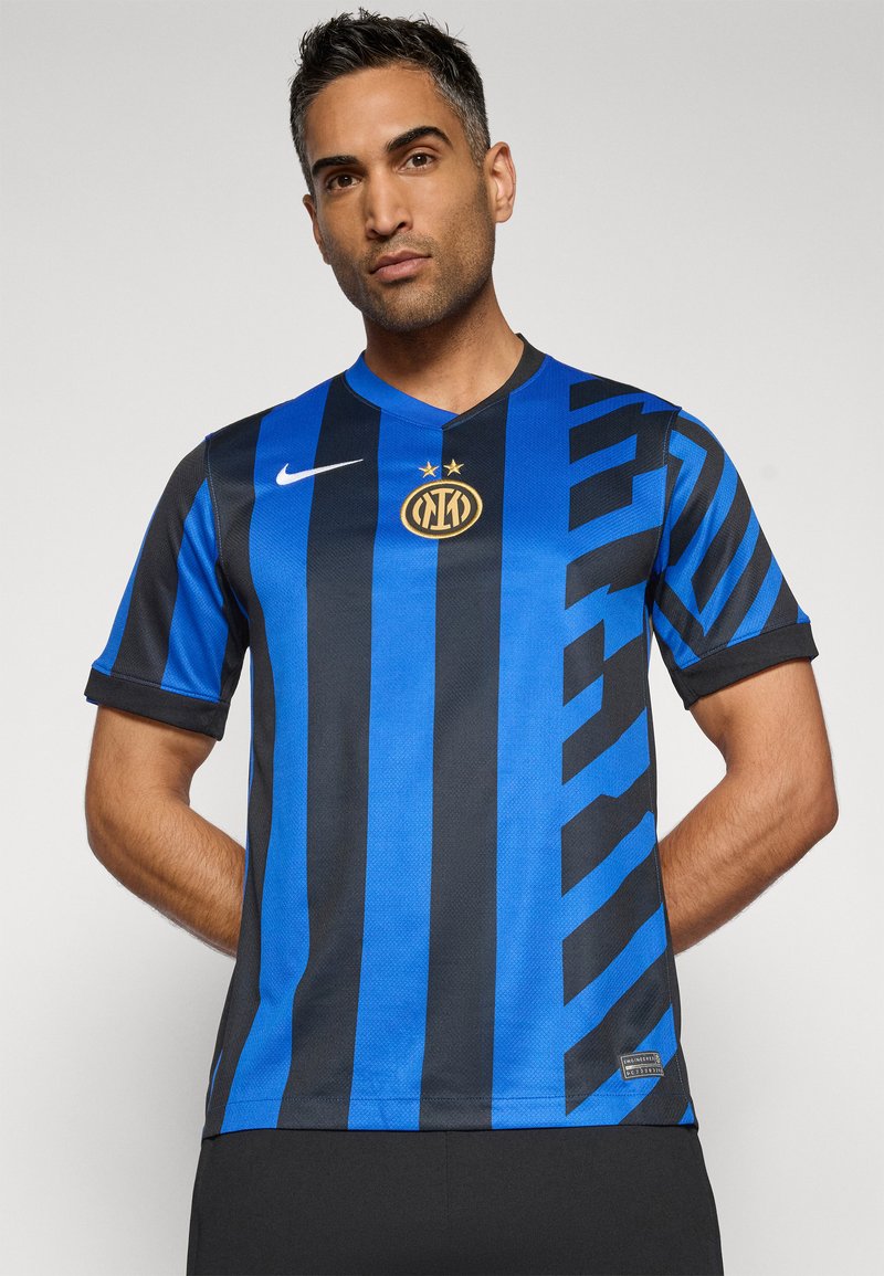 Hombre vistiendo una camiseta de fútbol del Inter de Milán a rayas negras y azules, con el logo del club en dorado y el swoosh de Nike, de pie con las manos detrás de la espalda.
