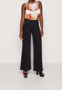 Czarne spodnie dzwony z wysokim stanem, w połączeniu z kolorową, wzorzystą koszulką bez rękawów. Modelka nosi czarne platformy.