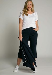 Witte korte mouwen V-hals T-shirt, zwarte cropped broeken, een marineblauwe jas vasthoudend en witte sneakers dragend. Eenvoudig ontwerp, casual pasvorm.