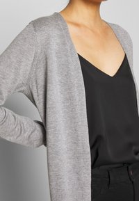Cardigan grigio in morbido materiale a maglia, design aperto davanti, con maniche lunghe. Indossato sopra un top nero a scollo a V, che mostra un look casual a strati.