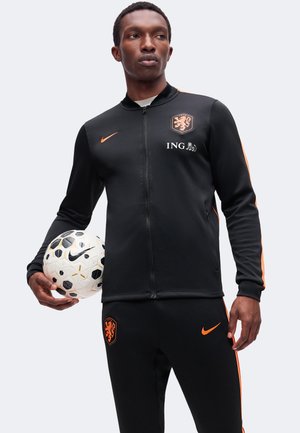 Mand i sort og orange sportstøj, der holder en hvid fodbold med sorte og gyldne detaljer og kigger til siden.