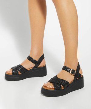 Jambes portant des sandales plateformes tissées noires avec des brides à la cheville et une boucle dorée, debout sur une surface blanche.