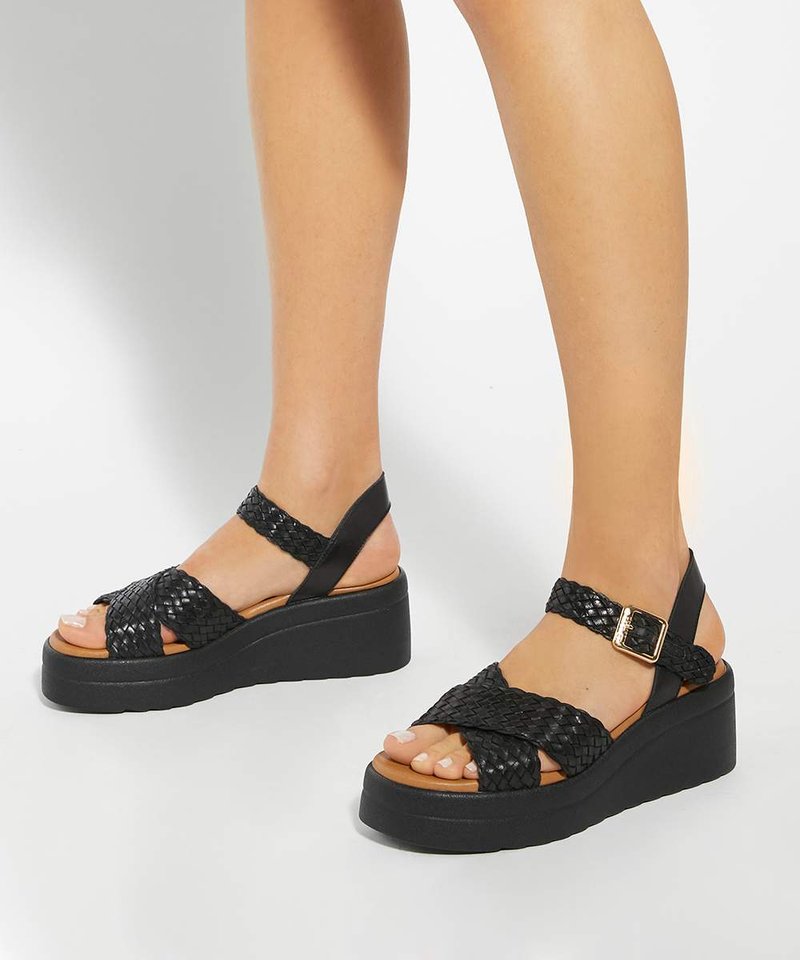 Jambes portant des sandales plateformes tissées noires avec des brides à la cheville et une boucle dorée, debout sur une surface blanche.