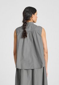 Blusa gris sin mangas con cuello, hecha de una tela ligera. Cuenta con un detalle fruncido en la espalda y un ajuste relajado, con un dobladillo ligeramente curvado.