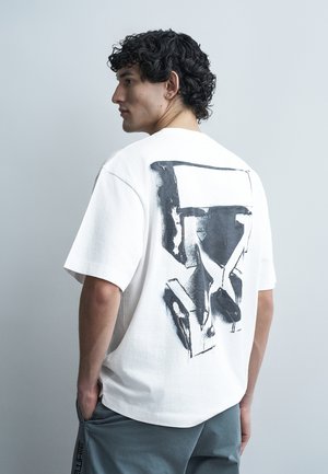 Νεαρός άνδρας με σγουρά μαλλιά, φορώντας λευκό φαρδύ T-shirt με αφηρημένο μαύρο και γκρι γεωμετρικό σχέδιο στην πλάτη, με τα χέρια στις τσέπες.