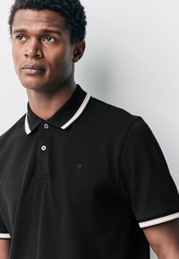 Musta poolopaita, jossa on kaksinappinen nappilista, joustinneuloskaulus, jossa on vaaleanruskeat raidat, ja hillitty kirjailtu logo rinnassa.