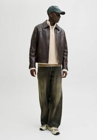 Homme portant une veste en cuir noir, un pull en tricot beige, un jean large délavé, des baskets blanches et une casquette de baseball vert foncé, debout et regardant à droite.