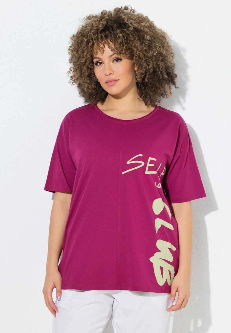 Femme aux cheveux bouclés portant un t-shirt ample magenta avec un texte crème et un pantalon blanc, debout devant un fond blanc.
