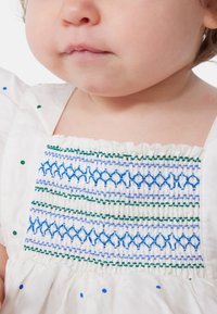 Abito bianco con intricate ricami blu e verde sul corpetto, rifinito con ruches arricciate lungo il collo e colorati pois.