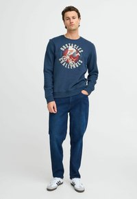 Mornarska dukserica s grafičkim printom, ležeran kroj, rebrasti završeci i porub. Uparen s plavim trapericama i bijelim tenisicama s crnim prugama.