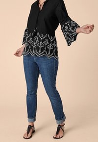 Blouse noire à manches cloche, ornée de motifs brodés blancs à l'ourlet. Portée avec un jean skinny bleu et des sandales noires.