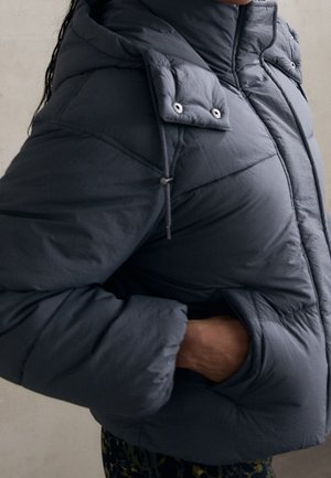 Persoon die een donker gewatteerde winterjas met hoge kraag draagt en hand in de jaszak tegen een effen achtergrond.