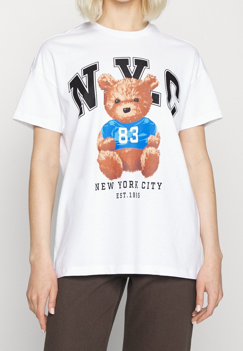T-shirt blanc avec un motif d'un ours en peluche marron portant un t-shirt bleu avec le numéro 83, avec "NYC" imprimé au-dessus et "New York City" en dessous.