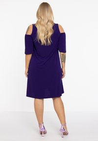 Lila Kurzarmkleid mit offenen Schultern, aus weichem Material. Hat einen lockeren Schnitt und einen abgerundeten Saum. Kombiniert mit helllila Absätzen.