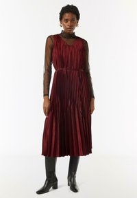 Burgunderfarbene, plissierte Midikleid mit V-Ausschnitt, darüber ein schwarzes, durchsichtiges Langarmtop. Getragen mit schwarzen, kniehohen Stiefeln mit Absatz.