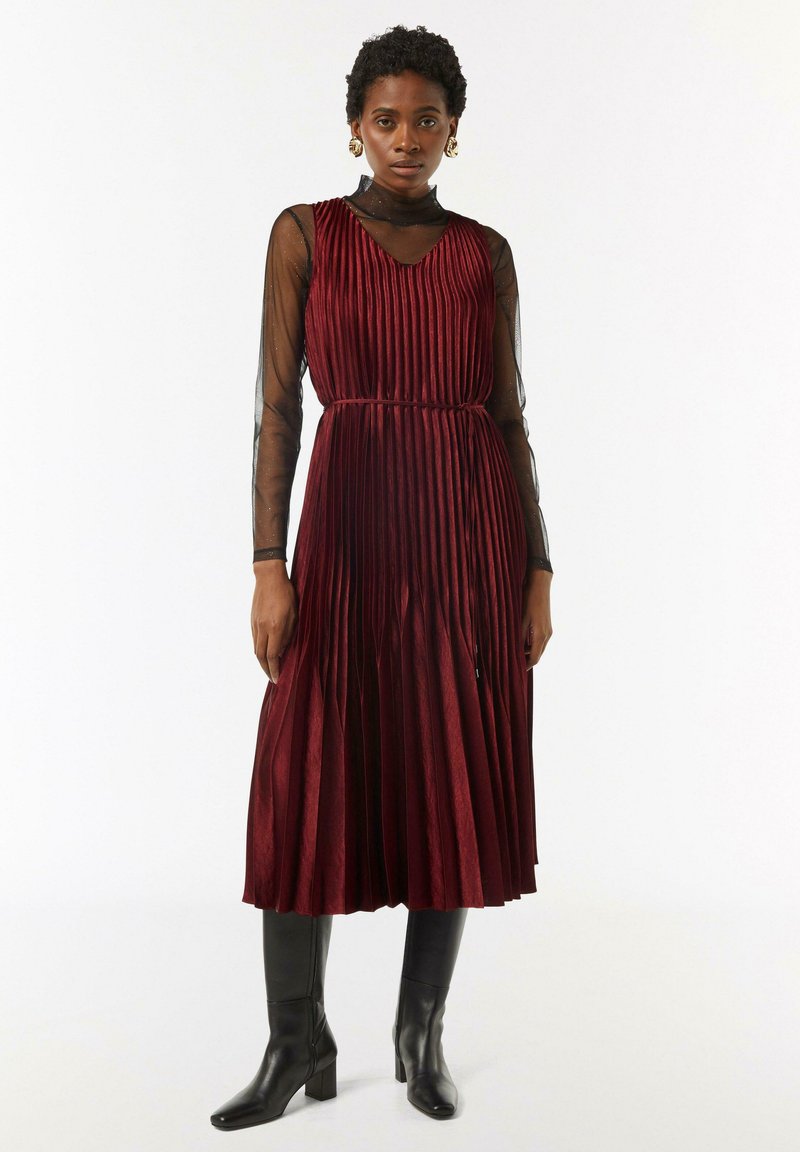 Burgunderfarbene, plissierte Midikleid mit V-Ausschnitt, darüber ein schwarzes, durchsichtiges Langarmtop. Getragen mit schwarzen, kniehohen Stiefeln mit Absatz.