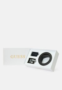 Ceinture en cuir noir avec des garnitures en argent, présentée dans une boîte blanche arborant le logo doré "GUESS". Comprend une boucle de ceinture rectangulaire.