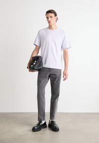 T-shirt mauve clair à manches courtes, jean gris tapered, mocassins noirs, tenant un sac noir. Tissu lisse, coupe décontractée, design minimaliste.