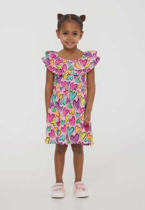 Abito colorato a motivi a cuore con sfondo rosa, maniche a volant e vita cinte. La ragazza indossa sneaker rosa chiaro con dettagli pastello.