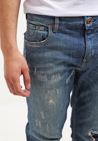 Los vaqueros de mezclilla azul claro presentan un corte regular, un ligero desgaste tipo whiskering y detalles desgastados en las rodillas y el muslo. Diseño de cinco bolsillos.