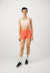 Sujetador deportivo rosa claro con un logo blanco, combinado con shorts naranjas de talle alto. La modelo lleva zapatillas deportivas negras y calcetines blancos hasta el tobillo.