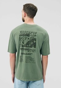 Olivegrünes, übergroßes T-Shirt mit einem grafischen Druck auf der Rückseite, der Konzertdetails zeigt, mit einem verblassten Look und doppelt genähten Säumen.