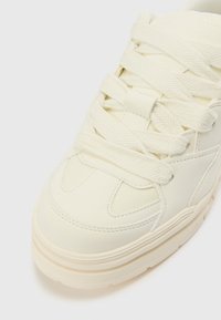 White leather sneaker with textured accents, thick laces, padded collar, and a chunky sole featuring subtle ridges for grip.