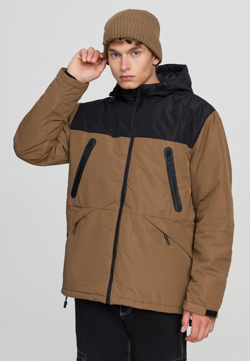 Brązowa i czarna puchowa kurtka z kapturem, z przednimi zamekami i bocznymi kieszeniami, charakteryzująca się gładką teksturą i regulowanymi sznurkami u dołu.