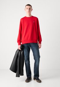Rød sweatshirt med ribbede manchetter og kant, båret med blå jeans og en sort læderjakke hængende over armen. Mørke sneakers fuldender looket.
