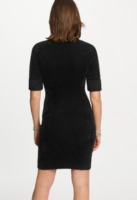 Donna con indosso un vestito corto nero a maniche pelose, in piedi con la schiena rivolta, con braccialetti su entrambi i polsi, su uno sfondo semplice.