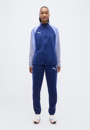 Nő áll előre nézve, kék és világoslila Puma melegítőben, fehér Puma logóval és fehér sportcipőben.