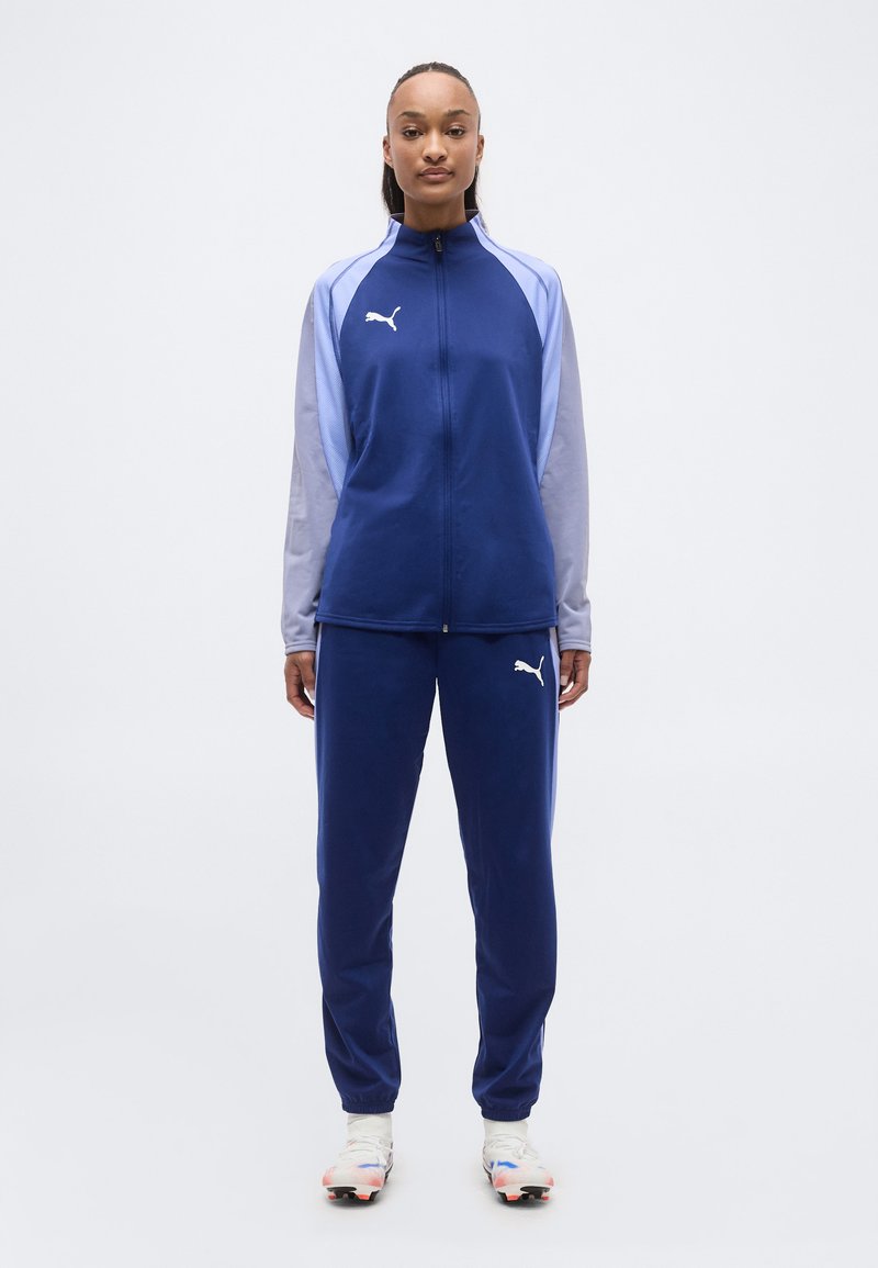 Γυναίκα που στέκεται και κοιτάζει μπροστά, φορώντας μπλε και ανοιχτό μοβ φόρμα Puma με λευκό λογότυπο Puma και λευκά αθλητικά παπούτσια.