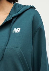 Približen posnetek osebe, ki nosi turkizen pulover z zadrgo, z belim logotipom New Balance na prsih in vrvicami na kapuci.