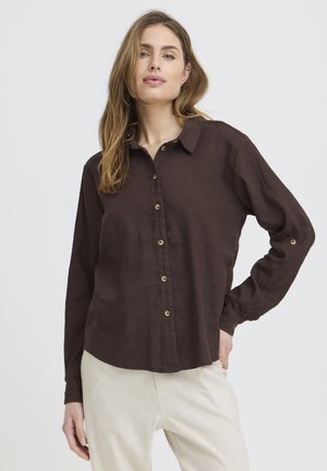Femme aux cheveux châtain clair portant une chemise à manches longues marron foncé boutonnée et un pantalon beige clair, debout avec une main sur la hanche.