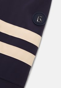 Granatowa tkanina z dwoma kremowymi poziomymi paskami. Posiada okrągłą granatową naszywkę z stylizowaną literą „B” w srebrnym wykończeniu.