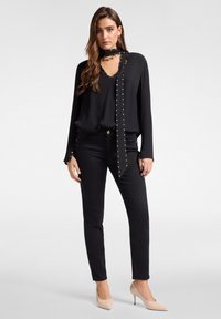 Mustapaita, jossa on V-pääntie ja pitkät hihat, jotka on korostettu kultaisilla niiteillä. Yhdistetty mustiin kapeisiin farkkuihin ja nude korkokenkiin.