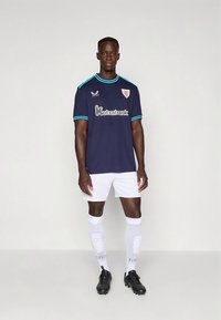 Marineblå fotballtrøye med lyseblå detaljer, med en logo, kombinert med hvite shorts og knelange sokker, brukt med svarte fotballsko.