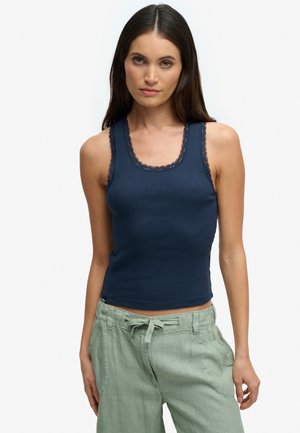 Kvinde iført en marineblå ribstrikket tanktop med blonder og lysegrønne bukser med snøre, stående mod en ensfarvet hvid baggrund.