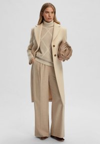 Cappotto di lana beige con grandi bottoni e spacco, indossato sopra un maglione chiaro con motivo a diamante e ampi pantaloni beige. La borsa ha una texture.