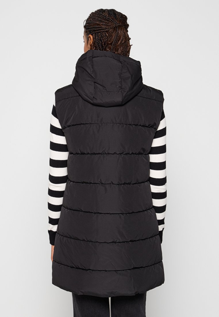 Giubbotto imbottito nero con cappuccio, caratterizzato da sezioni a quadrante orizzontale e una lunghezza maggiore, indossato sopra una maglietta a maniche lunghe a righe bianco e nero.