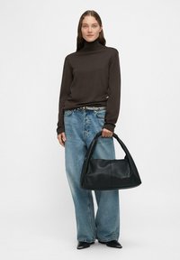 Czarna skórzana torebka o zaokrąglonym kształcie, trzymana przez osobę w brązowym golfie i jasnoniebieskich szerokich spodniach. Gładka faktura.