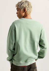 Felpa verde realizzata in tessuto morbido, con una vestibilità rilassata, polsini e orlo a coste. Dettaglio della cucitura posteriore visibile e design oversize.