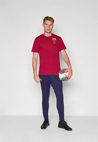 Vīrietis, kurš valkā sarkanu FC Barcelona T-kreklu un zilos bikses, tur rokās futbola bumbu ar melniem un oranžiem rakstiem.